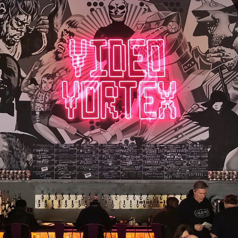 A glowing neon sign designed in a classic pixel aesthetic announces "Video Vortex" over a back bar.