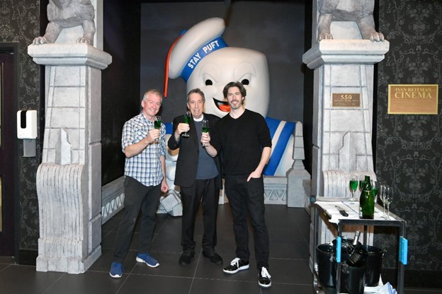 Ivan and Jason Reitman stand next to a Ghostbusters character outside a movie theater lobby.